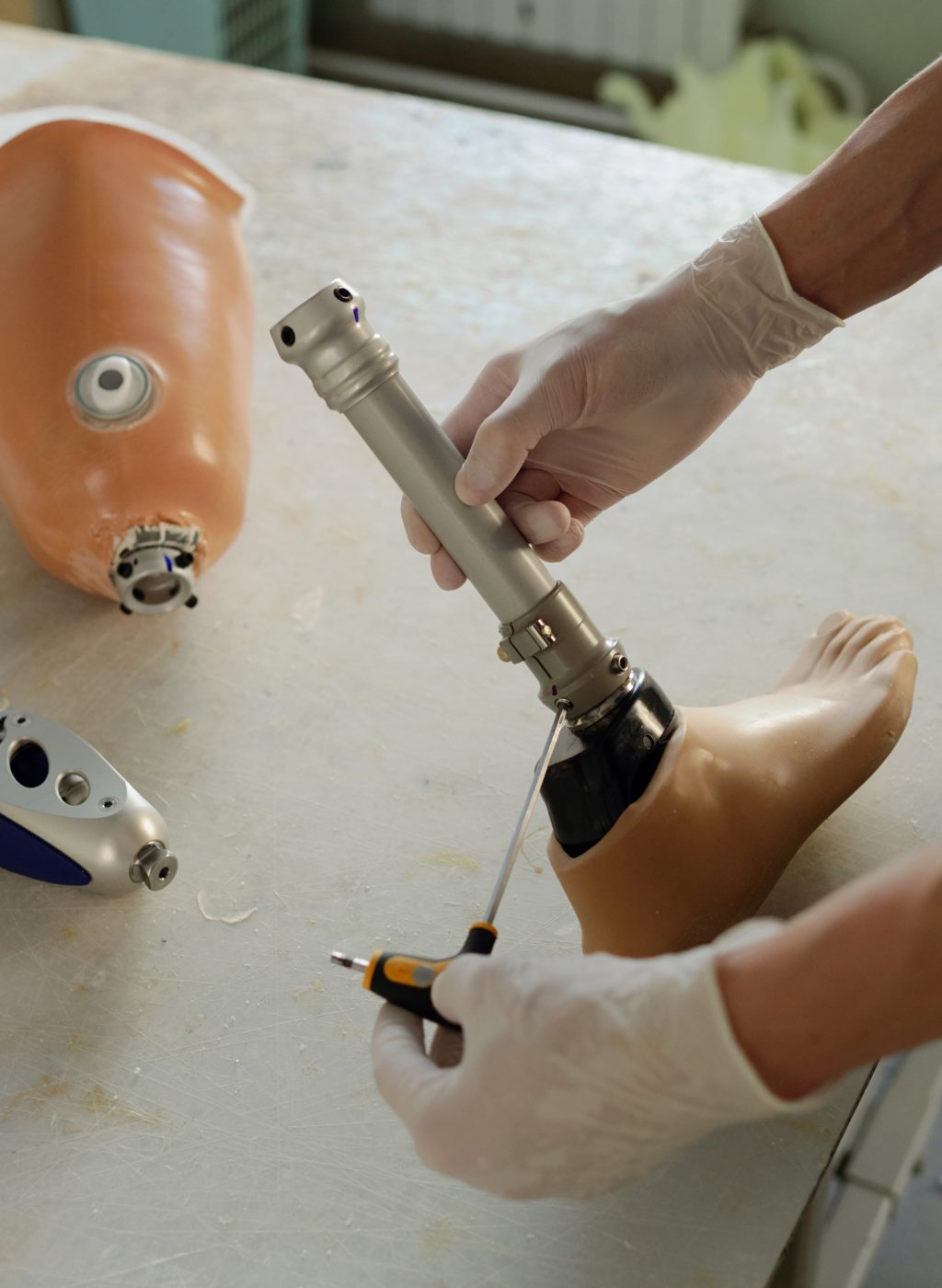 Hands of gloved manual worker of factory holding screwdriver and part of leg prosthesis over table while fixing steel shaft